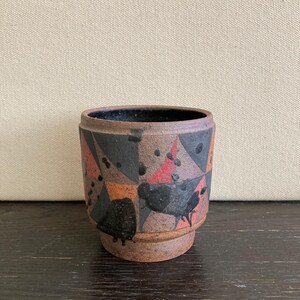 May include: A small, handcrafted ceramic pot with a geometric design. The pot features a brown base and rim, with a pattern of black, red, and orange shapes. The interior is dark. The pot is likely used for holding small items or as a decorative piece.