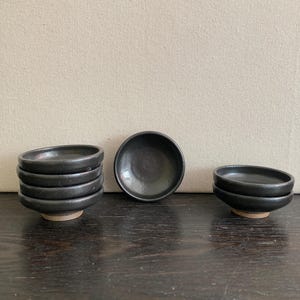 May include: A collection of small, black ceramic bowls. Several are stacked, while others are arranged individually. The bowls have a simple, minimalist design with a slightly rough, unglazed base. The dark color and matte finish give them a modern aesthetic.