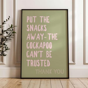 May include: A framed art print with a black border. The print has a light green background with pink text that reads "PUT THE SNACKS AWAY - THE COCKAPOO CAN'T BE TRUSTED THANK YOU."