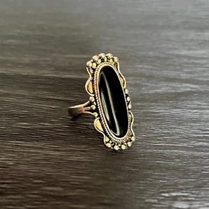 Peut inclure: Bague en argent ornée d'une pierre noire ovale. La bague présente un sertissage décoratif et orné avec de petits accents ronds autour de la pierre. La bague est posée sur un fond de grain de bois foncé.