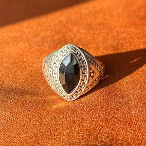 Puede incluir: Anillo plateado con una piedra preciosa negra ovalada. El anillo presenta una banda adornada con un diseño geométrico alrededor de la piedra preciosa. El anillo está sobre un fondo naranja, con una sombra proyectada a la derecha.
