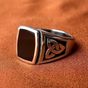Può includere: Un anello argentato con una pietra nera rettangolare e un motivo a nodo celtico. L'anello ha una finitura lucida e una fascia spessa. Il nodo celtico è in un quadrato incassato sul lato dell'anello.