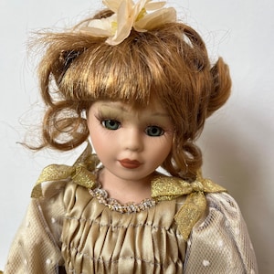Op de afbeelding: Een porseleinen pop met rossig haar en groene ogen. De pop draagt een gouden jurk met witte stippen en gouden strikken. Een bloem siert het haar van de pop. De pop heeft een serene uitdrukking.