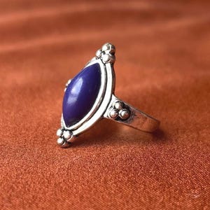 Könnte beinhalten: Ein Silberring mit einem marquiseförmigen, glatten, tiefvioletten Stein in einer Metallfassung. Der Ring und die Fassung sind mit kleinen, erhabenen Silberkugeln an der Oberseite, Unterseite und an den Seiten des Steins verziert.