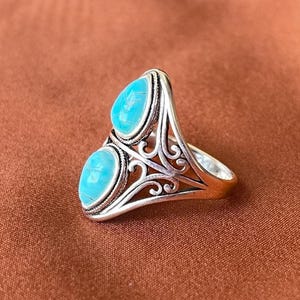 Peut inclure: Une bague en argent ornée de deux pierres bleues claires de forme ovale. La bague présente un motif ajouré orné de volutes. Les pierres sont serties dans un sertissage à lunette. La bague est sur une surface marron.