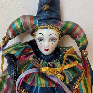 Può includere: Bambola di giullare decorativa in porcellana con cappello di raso blu e costume a righe multicolore. La bambola ha un viso dipinto con occhi blu, labbra rosse e un bordo dorato. Il cappello del giullare ha una piccola campana.