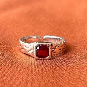 Peut inclure: Une bague ouverte de couleur argentée avec une pierre précieuse rouge foncé de taille carrée au centre. L'anneau en métal présente une finition martelée et texturée sur toute sa surface, et sa conception ouverte permet un ajustement réglable.