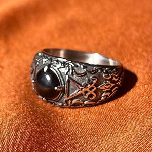 Könnte beinhalten: Ein silberfarbener Metallring mit einem polierten, schwarzen, runden Stein in der Mitte. Die Ringschiene ist mit komplexen, erhabenen Reliefmustern und einem dreieckigen Symbol verziert und weist eine texturierte, dunkle Oberfläche auf.