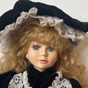 May include: A porcelain doll with blonde, curly hair and blue eyes. The doll wears a black velvet hat with white lace trim and a black velvet dress with a white lace collar. The doll's face is painted with delicate features.