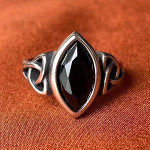 Op de afbeelding: Een zilveren ring met een centrale zwarte edelsteen in marquise-slijpvorm, gezet in een gepolijst metalen frame. De band toont aan beide zijden complexe opengewerkte knoopmotieven die uitlopen vanaf de centrale zetting.