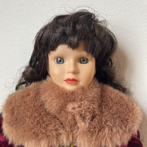 May include: A porcelain doll with dark brown hair and blue eyes. The doll wears a burgundy velvet dress with a faux fur collar. The dress is accented with gold trim and small gold beads.