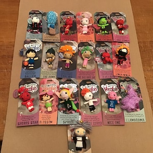 May include: A selection of Watchover Voodoo dolls, each with a distinct design and name, presented on their original packaging. The dolls showcase diverse colours and styles, including a ballerina, ninja, and sports star. The packaging includes text such as "Groove Mover" and "Nice One."