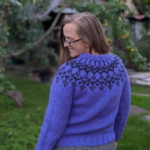 Peut inclure: Un pull en maille de couleur violette avec un motif géométrique noir et blanc sur l'épaule. Le pull est fait d'un fil doux et moelleux.