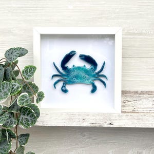 May include: A framed, blue crab art piece. The crab is a vibrant shade of turquoise with darker blue claws and legs. The artwork is set against a white background within a white square frame, creating a striking contrast. The crab art is a decorative piece.