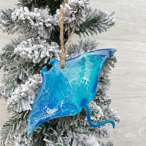 May include: A blue and white resin manta ray Christmas tree ornament, with a jute string. The ornament is displayed on a snow-covered Christmas tree. The manta ray has a glossy finish and a detailed design.
