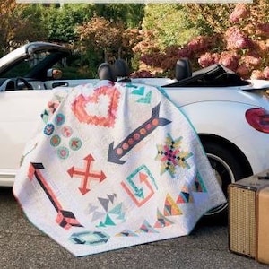 May include: A white quilt featuring a range of colourful patterns, including arrows, hearts, and geometric shapes. The quilt is displayed on the back of a white convertible car. The title "Follow Your Heart" and the text "10 Fun-To-Make Quilts You'll Love" are visible on the cover of a book in the background.