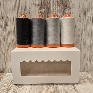 May include: Four spools of thread in a white box with a window. The spools are black, gray, light gray, and white. The spools have orange tops.