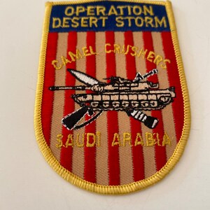 May include: A tan and red embroidered patch with a yellow border. The patch features a tank with crossed rifles and the text "Operation Desert Storm", "Camel Crushers", and "Saudi Arabia".