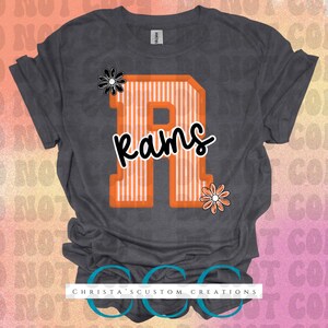 May include: Dark gray t-shirt featuring a large orange letter "R" with white stripes and the word "Rams" in a script font. The design includes two floral accents. The shirt has a crew neck and short sleeves.