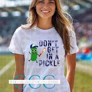 May include: White t-shirt with the phrase "DON'T GET IN A PICKLE" in navy blue text. The shirt features a cartoon pickle wearing a baseball cap and holding a baseball bat. The shirt has a crew neck and short sleeves.
