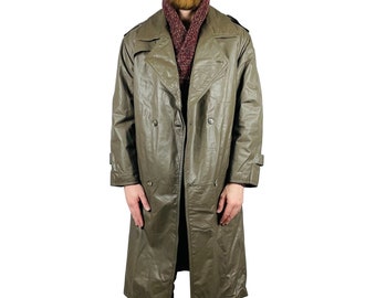May include: A long, olive green, double-breasted trench coat with a button closure. The coat has a collar and a belt. The model is wearing a gray beanie and a burgundy scarf.