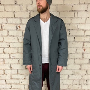 May include: A long, dark gray coat with a button closure and a collar. The coat has a relaxed fit and is made of a water-resistant material.