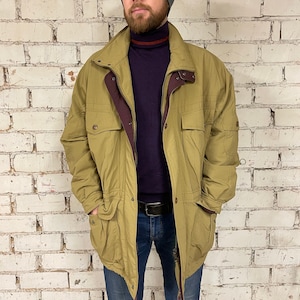 Puede incluir: Una chaqueta verde kaki con forro marrón, cierre de cremallera y varios bolsillos. La chaqueta tiene cintura con cordón y capucha.