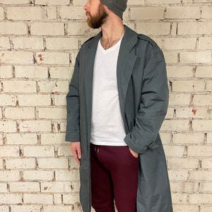 May include: A long, dark gray coat with a belt and a v-neck collar. The coat is worn over a white t-shirt and maroon sweatpants. The person is wearing a gray beanie.