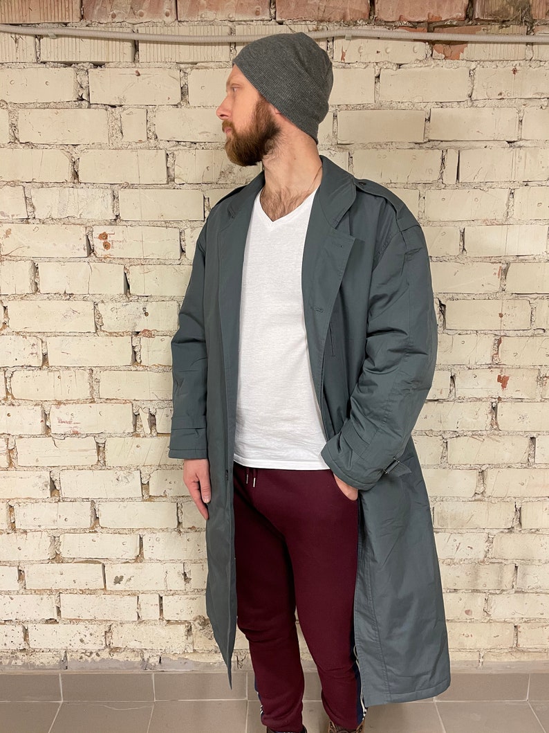 May include: A long, dark gray coat with a belt and a v-neck collar. The coat is worn over a white t-shirt and maroon sweatpants. The person is wearing a gray beanie.