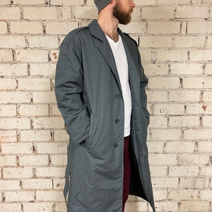 May include: A long, dark gray coat with a belt, a button closure, and two pockets. The coat is lined with a dark fabric. The person wearing the coat is standing in front of a white brick wall.