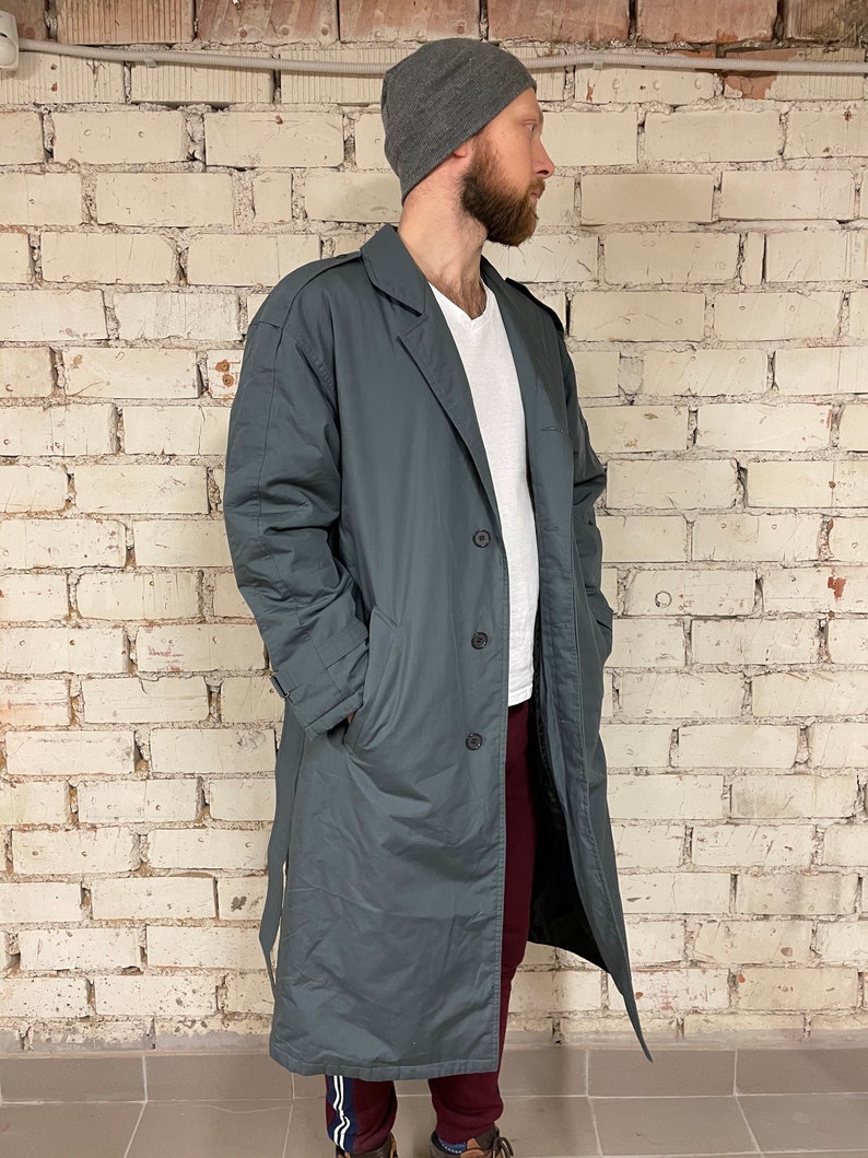 May include: A long, dark gray coat with a belt, a button closure, and two pockets. The coat is lined with a dark fabric. The person wearing the coat is standing in front of a white brick wall.