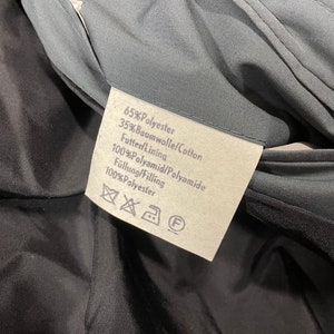 May include: A white fabric label with black text detailing the material composition of a garment. The label reads: 65% Polyester, 35% Baumwolle/Cotton, Futter Lining 100% Polyamid/Polyamide, F&uuml;llung/Filling 100% Polyester.
