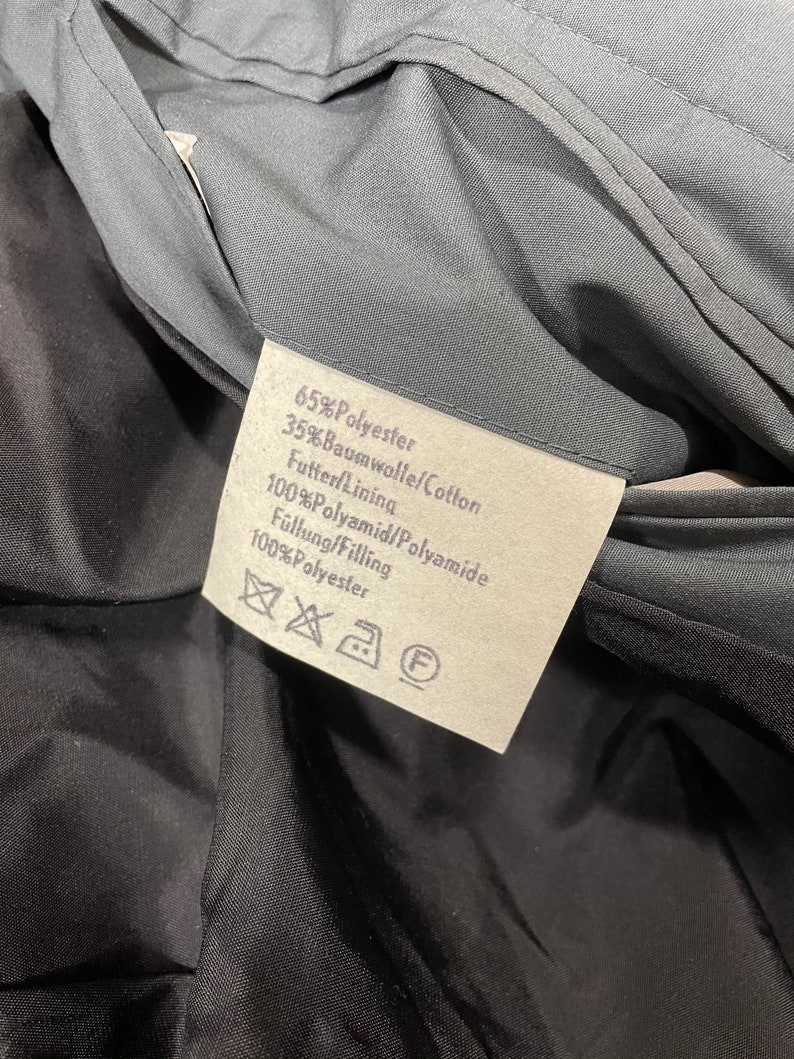 May include: A white fabric label with black text detailing the material composition of a garment. The label reads: 65% Polyester, 35% Baumwolle/Cotton, Futter Lining 100% Polyamid/Polyamide, F&uuml;llung/Filling 100% Polyester.