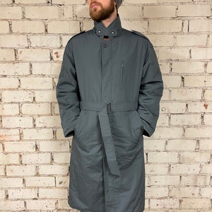 May include: A long, dark gray coat with a belt, a high collar, and a zipper closure. The coat has a single pocket on the left side and two pockets on the front.