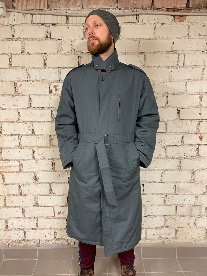May include: A long, dark gray coat with a belt, a high collar, and a zipper closure. The coat has a single pocket on the left side and two pockets on the front.