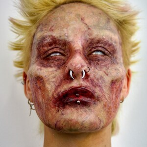 Puede incluir: Primer plano de la cara de una persona con maquillaje de zombie. La persona tiene el pelo rubio, los ojos blancos y la boca ensangrentada. Tiene varios piercings en la cara, incluyendo un aro en la nariz y un aro en el labio.