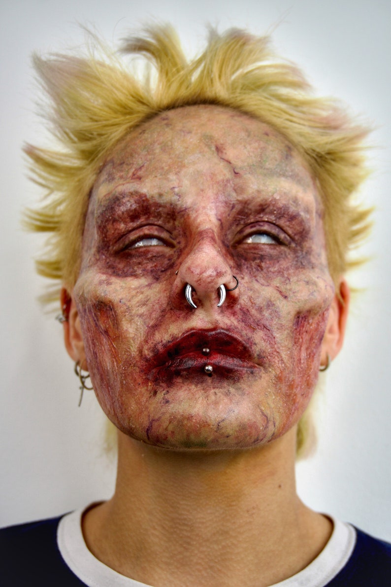 Puede incluir: Primer plano de la cara de una persona con maquillaje de zombie. La persona tiene el pelo rubio, los ojos blancos y la boca ensangrentada. Tiene varios piercings en la cara, incluyendo un aro en la nariz y un aro en el labio.