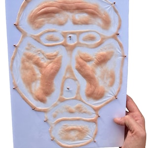 Può includere: Un volto protesico color carne, con dettagli precisi, è fissato a un supporto bianco. Spilli di rame fissano la protesi. Il volto comprende orbite oculari, naso e bocca, suggerendo un design realistico per effetti speciali o uso teatrale.