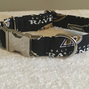 May include: Black dog collar featuring a purple and gold Baltimore Ravens logo. The collar has a silver buckle and a silver D-ring.