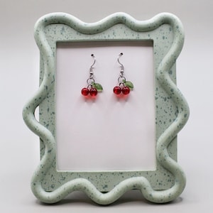 May include: A pair of cherry earrings with red beads and green leaf accents. The earrings hang from silver-colored hooks. The earrings are displayed in a light green, wavy-edged frame with a speckled pattern.