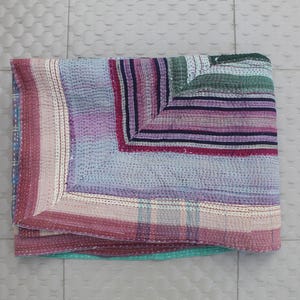 Puede incluir: Colcha plegada multicolor con diseño de patchwork. La colcha presenta una variedad de colores, incluyendo rosa, morado, azul y verde, dispuestos en rayas y cuadrados horizontales. Los bordes están acabados con un borde contrastante.