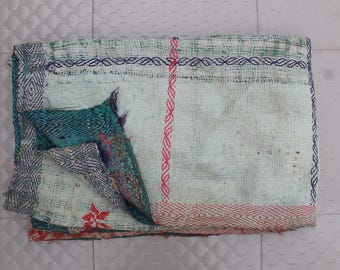 May include: A folded, vintage textile with a light blue and white base. The edges are detailed with dark blue stitching and geometric patterns. A section of teal fabric is visible, and a red floral design is present.