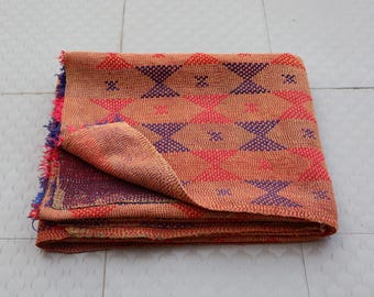 May include: A folded textile featuring a tan base with a repeating geometric pattern of red and blue triangles. The fabric displays visible running stitch details throughout the surface, with frayed, multi-coloured edges showing red and blue threads.