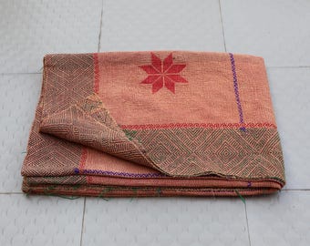 May include: A folded peach-colored textile featuring a red eight-pointed star motif in the center. The fabric displays a dark green diamond-patterned border along the edges, accented by thin blue and red stitched lines throughout the material.