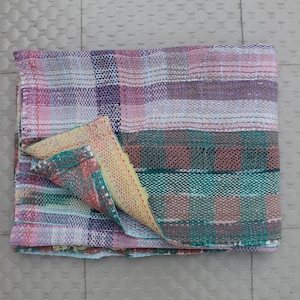 Puede incluir: Textil plegado multicolor con diseño patchwork. La manta presenta una mezcla de tonos rosa, morado, verde y blanco en un patrón tejido. La esquina está doblada, revelando un reverso amarillo contrastante y estampado.