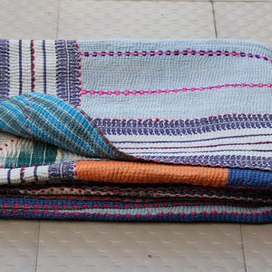 Op de afbeelding: Een gevouwen rechthoekige quilt met een lichtblauwe basis en horizontale strepen in donkerblauw, wit en roze. De stof vertoont overal ingewikkelde stikselpatronen, waarbij een hoek de groen en blauw gepatroonde onderkant laat zien.