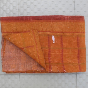 May include: A folded orange and red Kantha quilt, showing its textured surface. The quilt has a patchwork design with visible stitching and a folded corner, displaying the fabric's layers. The colour scheme is warm, with shades of orange and red.