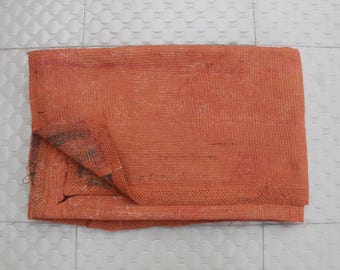 May include: An orange textile with a folded corner, showcasing a woven texture. The fabric appears to be a blanket or throw, with visible stitching and a slightly distressed appearance. The color is a warm, earthy tone.