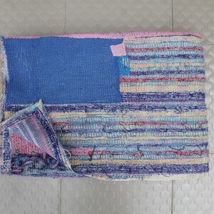 Puede incluir: Textil rectangular hecho a mano con un diseño de patchwork. La sección superior es azul liso, mientras que la sección inferior presenta rayas horizontales en tonos de azul, morado, rosa y amarillo. Una esquina está doblada, revelando el interior.