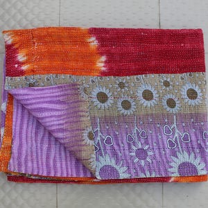 May include: A folded, multi-colored textile with a patchwork design. The top section is orange and red, transitioning to a floral and striped pattern in shades of purple, tan, and white. The textile is likely a blanket or quilt.
